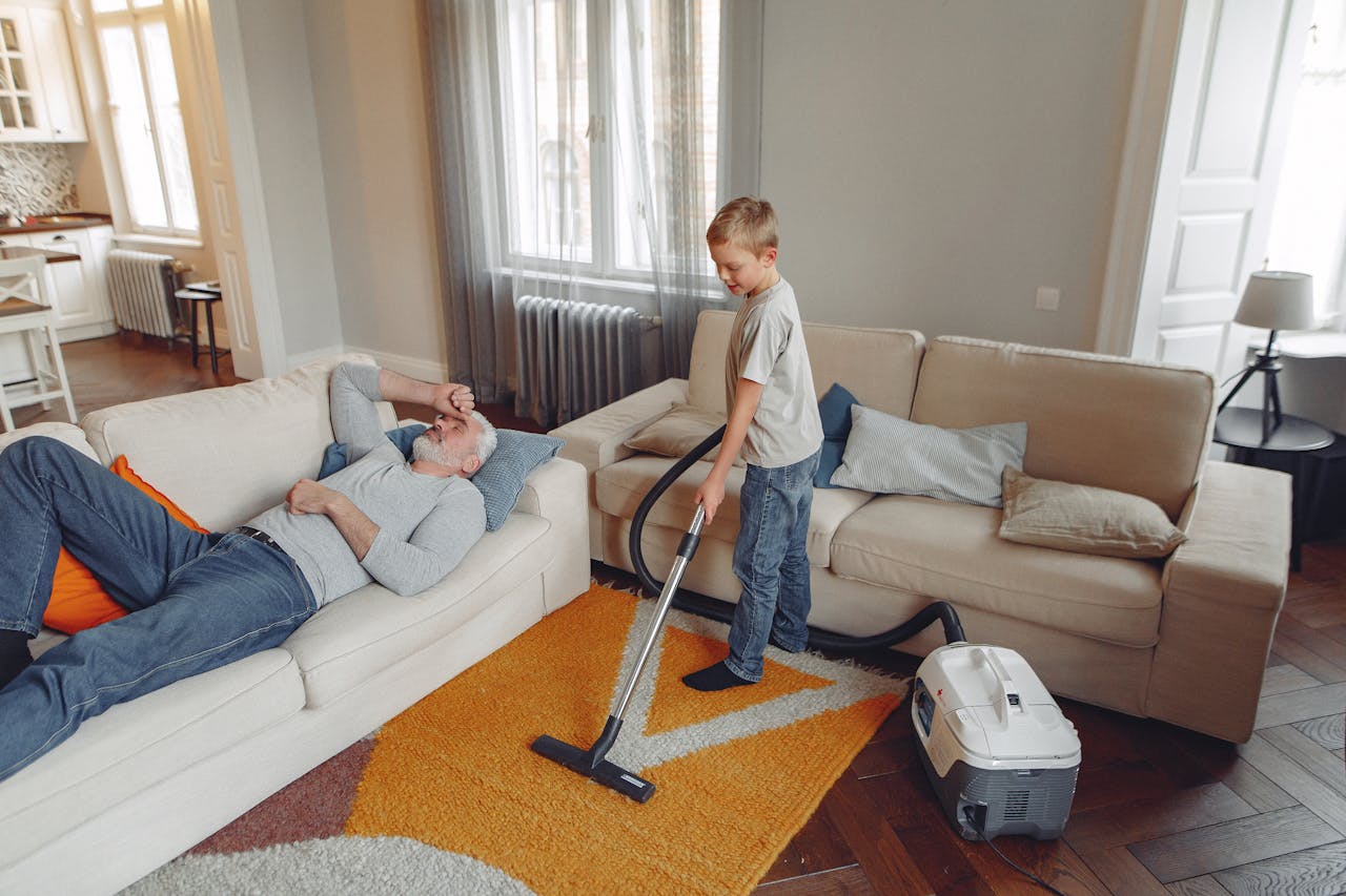 portfolio-02 A young boy vacuums the living room carpet while his grandfather relaxes on the sofa.