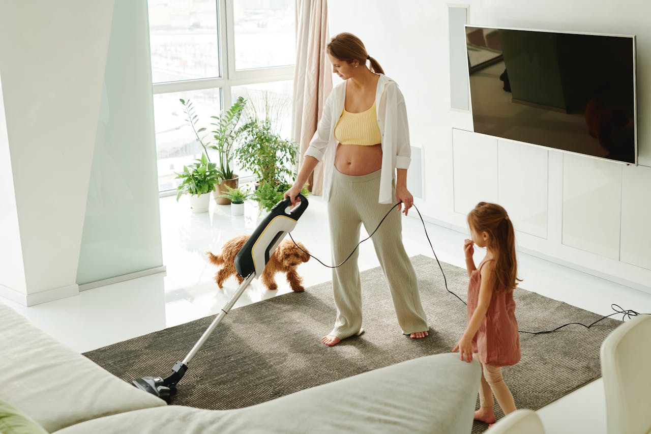 portfolio-04 Pregnant mother cleaning carpet with vacuum as daughter and dog play indoors.